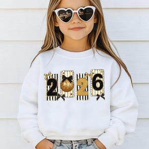 May include: White sweatshirt featuring the numbers "2026" in a black and gold design. The sweatshirt has a crew neck and long sleeves. The text "Tiny Baby Love" is at the top right.