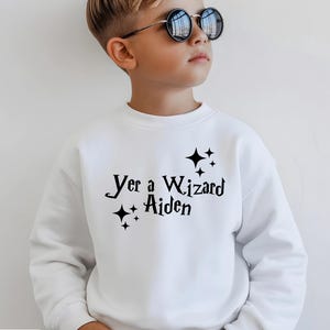 May include: White sweatshirt with the text "Yer a Wizard Aiden" in black script, accented with star graphics. The sweatshirt is a crew neck style. The word "WHITE" is in a white rectangle at the bottom left.