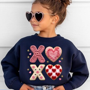 May include: A navy blue sweatshirt with the text "Tiny Baby Love" and the word "NAVY". The sweatshirt features embroidered "XO" designs with hearts and stars. The child is wearing heart-shaped sunglasses.