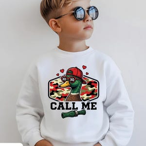 Può includere: Felpa bianca con grafica di un'anatra con occhiali da sole e berretto rosso, con la scritta "CALL ME". L'anatra è ambientata su uno sfondo mimetico. La felpa ha anche la scritta "WHITE" in basso.