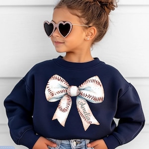 May include: Navy blue sweatshirt featuring a baseball-themed bow design. The bow is white with red stitching, resembling a baseball. The name "Amelia" is printed on one of the bow's tails. The text "Tiny Baby Love" is in the upper right corner.