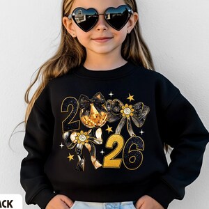 May include: Black sweatshirt with a gold and black graphic design featuring the numbers "2026" and bows. The sweatshirt has a crew neck and long sleeves. The word "BLACK" is printed at the bottom left.