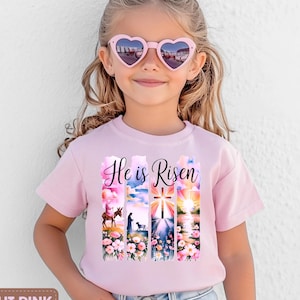 May include: A light pink t-shirt with the words "He is Risen" and a colourful graphic. The graphic shows scenes with a cross, flowers, and a sunrise. The t-shirt is worn by a young girl wearing heart-shaped sunglasses.