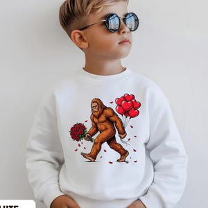 Puede incluir: Sudadera blanca con un dibujo animado de Pie Grande llevando rosas rojas y globos con forma de corazón. El texto "Tiny Baby Love" está en la parte superior derecha. La palabra "WHITE" está en la parte inferior izquierda.