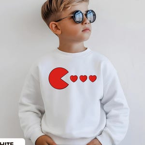 May include: White sweatshirt featuring a red Pac-Man-like design with three red hearts. The sweatshirt has the word "WHITE" printed at the bottom and the text "Tiny Baby Love" at the top. The sweatshirt is worn by a child.