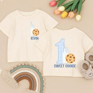 Könnte beinhalten: Zwei cremefarbene T-Shirts mit kurzen Ärmeln. Ein Shirt hat eine Milchflaschen- und Keks-Grafik mit dem Namen "KEVIN". Das andere Shirt hat eine blau karierte Zahl "1", eine Milchflasche und eine Keks-Grafik mit dem Text "SWEET COOKIE".