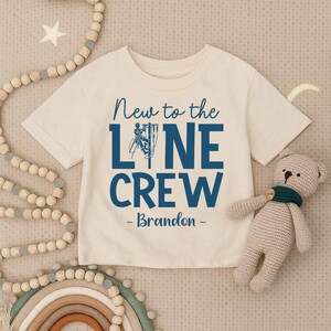 May include: A cream-colored short-sleeve t-shirt featuring dark blue text that reads New to the LINE CREW - Brandon - with a graphic of a utility worker on a pole replacing the letter I in the word LINE. The shirt has a crew neckline.