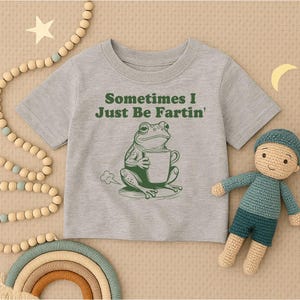以下が含まれることがあります： 「Sometimes I Just Be Fartin'」の文字とマグカップを持った緑色のカエルのイラストが描かれたグレーのTシャツ。人形、虹、ビーズガーランドと一緒に表示されています。