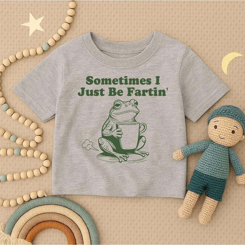以下が含まれることがあります： 「Sometimes I Just Be Fartin'」の文字とマグカップを持った緑色のカエルのイラストが描かれたグレーのTシャツ。人形、虹、ビーズガーランドと一緒に表示されています。