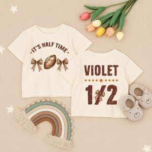 May include: Two cream-colored baby t-shirts. One shirt says "It's Half Time" with a football and bows. The other says "Violet 1/2" with stars. A rainbow wall hanging, baby shoes, and flowers are also in the image.