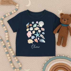 Peut inclure: T-shirt bleu marine avec une collection de coquillages et une étoile de mer à l'aquarelle. Le nom "Oliver" est imprimé en cursive. Un ours en peluche marron et des perles décoratives sont en arrière-plan.