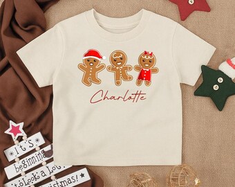 May include: A cream-colored t-shirt featuring three gingerbread people with festive hats and dresses. The name "Charlotte" is written in red script below the gingerbread figures. Christmas-themed decorations surround the shirt.