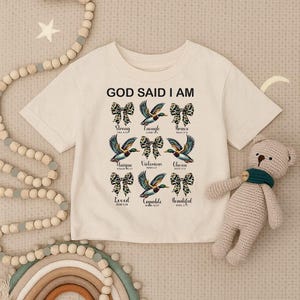 以下が含まれることがあります： 「GOD SAID I AM」の文字とリボンとアヒルのイラストが描かれたクリーム色のTシャツ。「Strong」「Enough」「Brave」などの言葉が特徴です。
