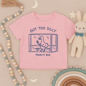 May include: A light pink short-sleeve t-shirt featuring a dark blue graphic of a duck holding a hockey stick inside a penalty box. The text above the graphic reads GOT TOO SILLY and the text below the graphic reads PENALTY BOX in dark blue.