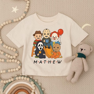 May include: Cream-colored short-sleeve t-shirt featuring a graphic of cartoon horror characters. The design includes Freddy Krueger, Jason Voorhees, Pennywise, Ghostface, and Sam. The text "MATTHEW" is printed below the characters.