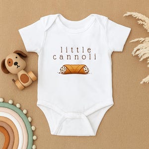 Pode incluir: Body de bebé branco com as palavras "little cannoli" em texto castanho sobre uma ilustração de um cannoli. O cannoli é castanho com recheio branco e castanho. O body está sobre uma superfície bege.