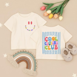 以下が含まれることがあります： クリーム色の半袖Tシャツ2枚。1枚は胸元にスマイリーフェイスの小さなグラフィックと「COOL COUSINS CLUB」の文字が入っています。もう1枚は、青い縦縞の上にスマイリーフェイスをあしらった、大きくカラフルな「COOL COUSINS CLUB」のグラフィックが描かれています。