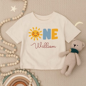 Op de afbeelding: Crèmekleurig T-shirt met een lachende zon-afbeelding en het woord "ONE" in blauw en geel. De naam "William" staat eronder in bordeauxrood schrift. Een knuffelbeer en houten kralen zijn op de achtergrond.
