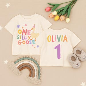 May include: Two cream-colored short-sleeve t-shirts. One shirt features a graphic of a goose wearing a party hat with the text "ONE SILLY GOOSE" and small flowers. The other shirt displays the name "OLIVIA" in colorful letters above the number "1".