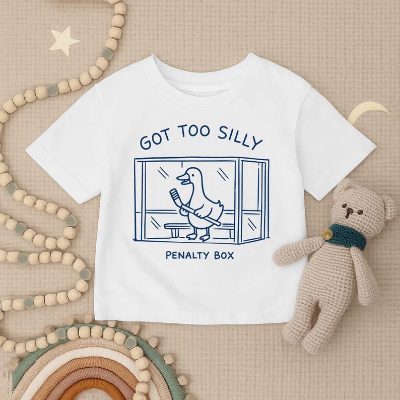 May include: A white short-sleeved t-shirt featuring a dark blue graphic of a duck holding a hockey stick inside a penalty box. The text above the graphic reads GOT TOO SILLY and the text below the graphic reads PENALTY BOX in dark blue ink.