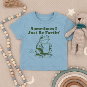 以下が含まれることがあります： 「Sometimes I Just Be Fartin'」の文字と、マグカップを持った緑色のカエルのイラストが描かれた水色のTシャツ。柔らかい素材でできており、子供向けです。