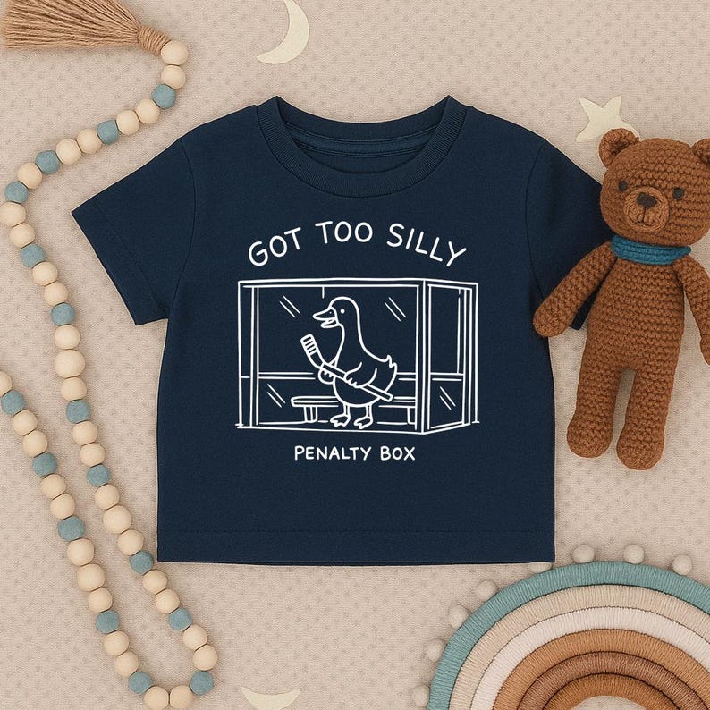 May include: A navy blue short-sleeve t-shirt featuring a white line-art graphic of a duck holding a hockey stick inside a penalty box. The text above the graphic reads GOT TOO SILLY and the text below the graphic reads PENALTY BOX.