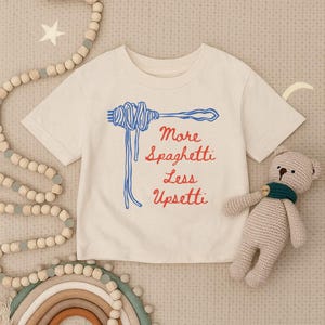May include: Off-white t-shirt with a blue spaghetti and fork graphic and the text "More Spaghetti Less Upsettti" in red. A small, beige teddy bear with a green scarf is next to the shirt.