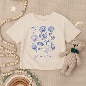 May include: A cream-colored short-sleeve t-shirt featuring a blue graphic print of various seashells, starfish, and small bows. The name Amelia is printed in blue script below the collection of sea-themed illustrations on the front of the shirt.
