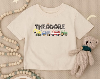 May include: A cream-coloured t-shirt featuring the name "THEODORE" in grey block letters. Below the name are colourful illustrations of construction vehicles: a yellow excavator, a blue truck, a cement mixer, a red fire engine, and a green tractor. A teddy bear is in the image.