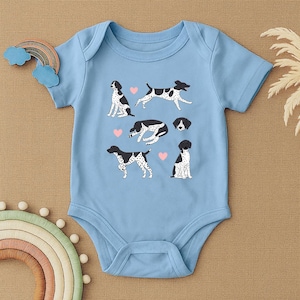 May include: A light blue baby bodysuit featuring illustrations of dogs in various poses, with black and white markings and pink heart accents. The bodysuit is made of soft fabric.