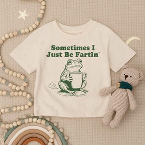 以下が含まれることがあります： 「Sometimes I Just Be Fartin'」の文字と、マグカップを持った緑色のカエルのイラストがプリントされたクリーム色のTシャツ。右側には、緑色のスカーフを巻いたベージュのぬいぐるみがあります。