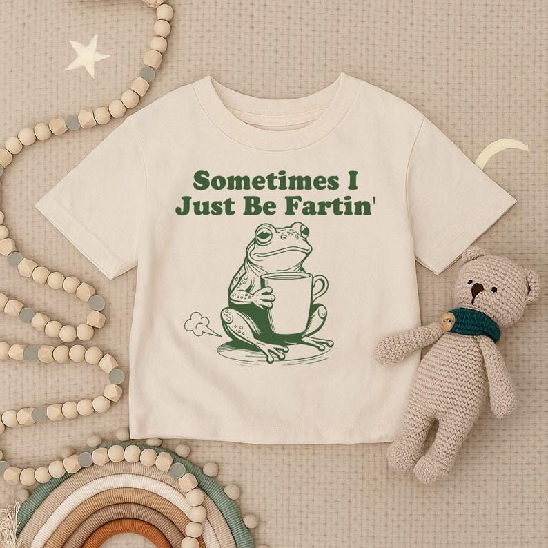 以下が含まれることがあります： 「Sometimes I Just Be Fartin'」の文字と、マグカップを持った緑色のカエルのイラストがプリントされたクリーム色のTシャツ。右側には、緑色のスカーフを巻いたベージュのぬいぐるみがあります。