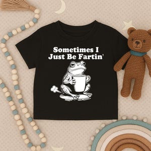 以下が含まれることがあります： 「Sometimes I Just Be Fartin'」の文字とマグカップを持ったカエルのグラフィックがプリントされた黒い子供用Tシャツ。背景には茶色のテディベアとビーズガーランドがあります。Tシャツは柔らかい素材で作られています。