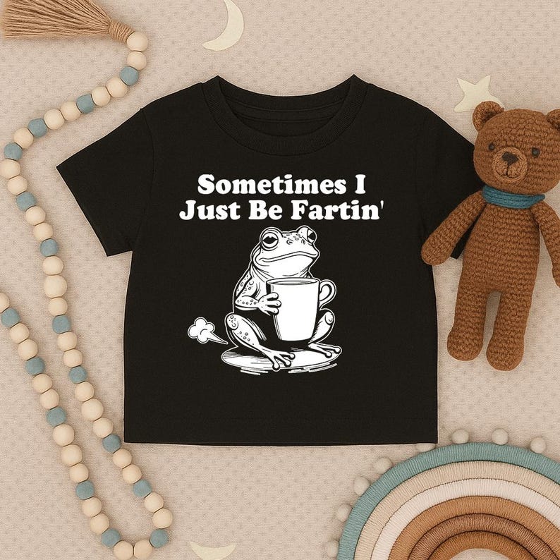 以下が含まれることがあります： 「Sometimes I Just Be Fartin'」の文字とマグカップを持ったカエルのグラフィックがプリントされた黒い子供用Tシャツ。背景には茶色のテディベアとビーズガーランドがあります。Tシャツは柔らかい素材で作られています。
