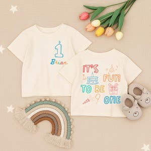 May include: Two cream-colored short-sleeve t-shirts. One features a blue number one and the name Brian. The other displays the text IT'S FUN TO BE ONE with illustrations of a cake, gift, balloon, and party hats in various colors.