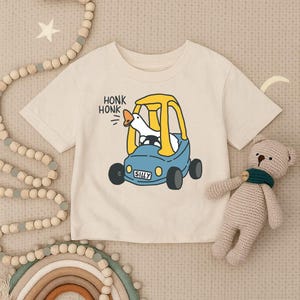 May include: Cream-colored short-sleeved t-shirt featuring a cartoon goose driving a blue car with yellow accents. The text "HONK HONK" and "SILLY" are printed on the shirt. A stuffed bear toy is next to the shirt.