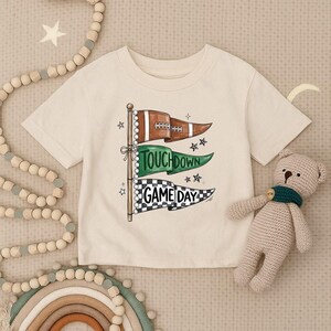 Könnte beinhalten: Ein cremefarbenes Kurzarm-T-Shirt mit einer Grafik aus drei übereinander angeordneten Wimpeln an einem Mast. Der obere Wimpel ist braun mit einem Football-Motiv, der mittlere ist grün mit dem Text TOUCHDOWN und der untere ist schwarz-weiß kariert.