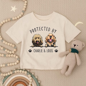 May include: Cream-colored t-shirt featuring the text "PROTECTED BY" above two dog illustrations. Below the dogs, the text reads "CHARLIE & LOUIS" with paw prints. A teddy bear and decorative beads are in the background.