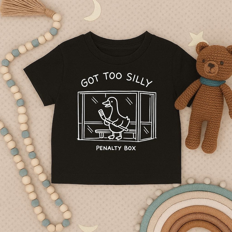 May include: A black short-sleeved t-shirt featuring a white line-art graphic of a duck holding a hockey stick inside a penalty box. The text above the graphic reads GOT TOO SILLY and the text below the graphic reads PENALTY BOX in white.