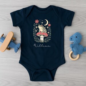 Pode incluir: Body de bebé azul marinho com uma ilustração de rã. O design apresenta uma rã num cogumelo, rodeada por flores e uma lua crescente. O nome "William" está escrito abaixo.