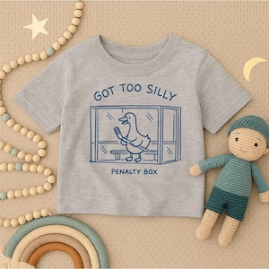 May include: A heather gray short-sleeve t-shirt featuring a dark blue graphic of a duck holding a hockey stick inside a penalty box. The text above the graphic reads GOT TOO SILLY and the text below the graphic reads PENALTY BOX.