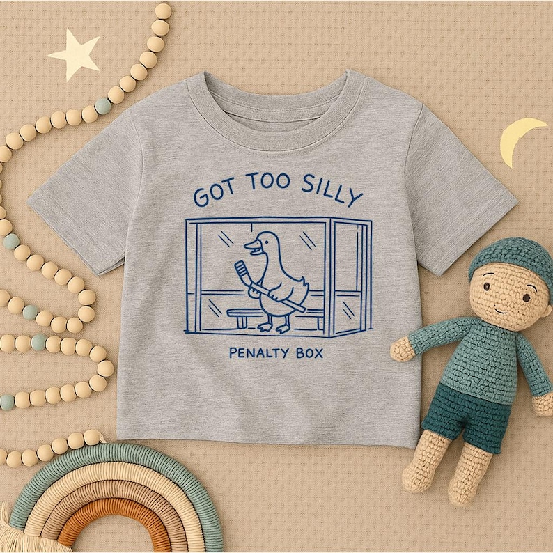 May include: A heather gray short-sleeve t-shirt featuring a dark blue graphic of a duck holding a hockey stick inside a penalty box. The text above the graphic reads GOT TOO SILLY and the text below the graphic reads PENALTY BOX.