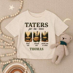May include: Off-white t-shirt featuring the words "TATERS po-ta-toes" and images of cooking potatoes. The shirt also has the words "boil em", "mash em", and "stick em a stew". The name "THOMAS" is printed below.