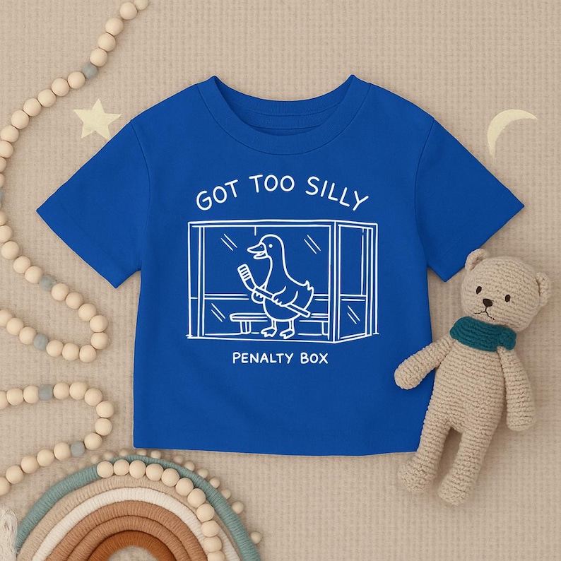 May include: A royal blue short-sleeve t-shirt featuring a white line art graphic of a duck holding a hockey stick inside a penalty box. The text above the graphic reads GOT TOO SILLY and the text below the graphic reads PENALTY BOX.