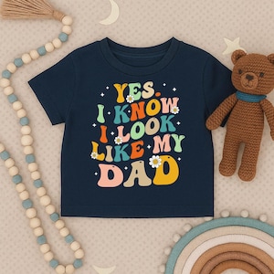May include: Navy blue toddler t-shirt with the text "YES, I KNOW I LOOK LIKE MY DAD" in colorful, retro-style lettering. The shirt features daisy accents and is next to a brown teddy bear.