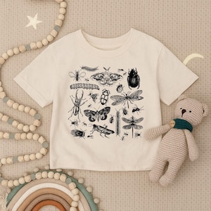 May include: A cream-colored short-sleeve t-shirt featuring a black ink-style graphic print of various insects, including beetles, moths, dragonflies, and centipedes, arranged in a central cluster on the front of the garment.