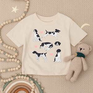 May include: Cream-colored t-shirt featuring a playful design of black and white dogs in various poses, with pink heart accents. The shirt is made of soft material and is suitable for children.