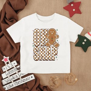May include: White toddler t-shirt with a brown and white checkered design that spells out "OH SNAP" with a gingerbread man graphic. The shirt is decorated with green and red accents, perfect for the holiday season.