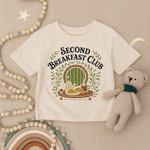 May include: A cream-colored short-sleeve t-shirt featuring a graphic print with the text "SECOND BREAKFAST CLUB" above an illustration of a round green door, a teapot, a mug, and croissants, all framed by green leafy branches.