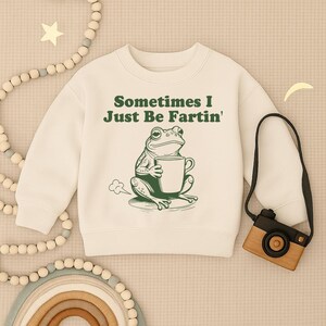 以下が含まれることがあります： 「Sometimes I Just Be Fartin'」の文字がプリントされたクリーム色のスウェットシャツ。緑色のカエルのイラストがマグカップを持っています。スウェットシャツはクルーネックと長袖です。カエルは雲の上に座っています。
